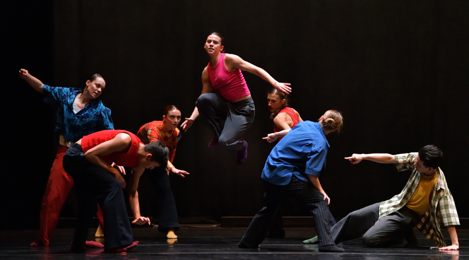 Ballet dancers in colouful urban, athletic clothing pose in the middle of a ballet performance with six members surrounding a woman in the centre who is mid jump.