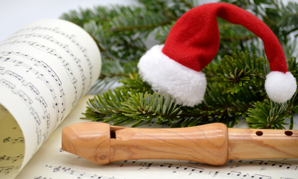 a tiny santa hat sits atop a pine tree branch next to a wooden recorder and sheet music.