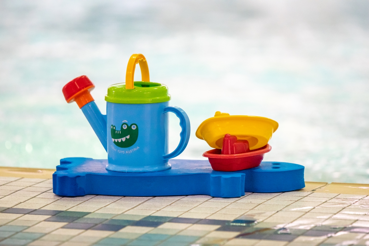 A toy watering can and small plastic boats sit on top of a blue turtle-shaped kickboard by the edge of the Winskill Teach Pool.