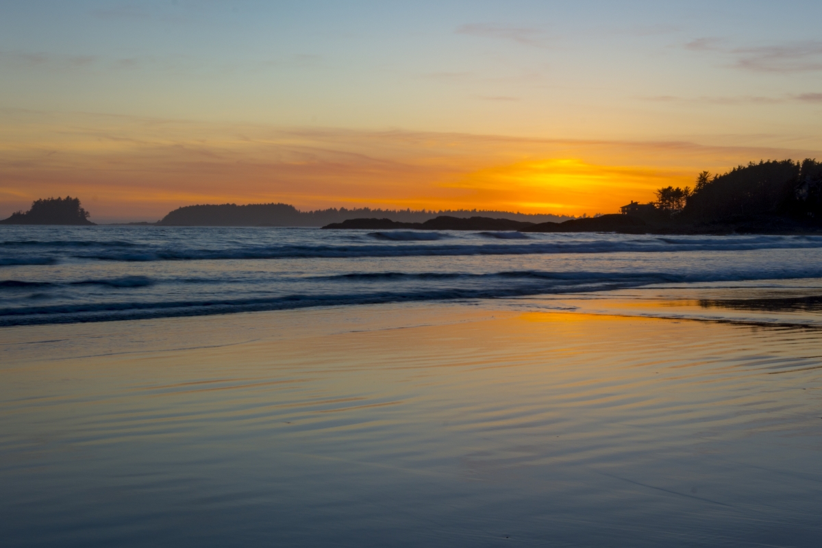Glowing yellow sun, setting behind the rocks, water, and sand of Tofino. 