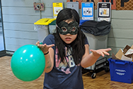 Girl in eye mask shrugs while holding a green balloon
