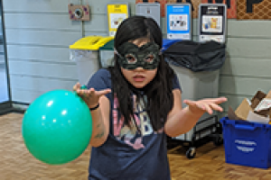 Girl in eye mask shrugs while holding a green balloon